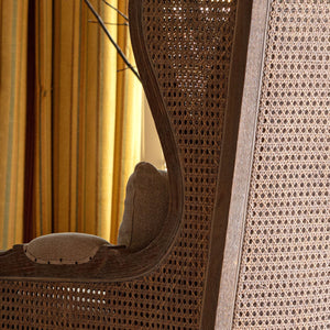 A close-up of a woven cane or rattan chair back and armrest in warm brown tones, with a beige upholstered seat cushion and a softly lit indoor background with yellow curtains.