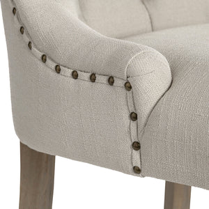 Close-up of a light beige upholstered armchair showing the fabric texture, rolled arm, nailhead trim, and part of the wooden leg against a white background.