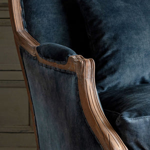Close-up of a dark blue velvet upholstered armchair with ornate carved wooden arms and a matching cushion.