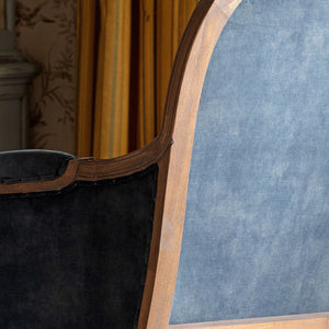 Close-up of an upholstered chair or sofa showing a blue fabric backrest, dark charcoal velvet seat, and curved wooden frame in an indoor setting with yellow curtains in the background.