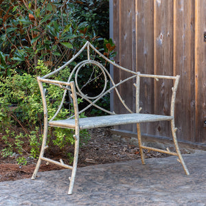A rustic garden bench made from branches and metal, featuring a circular center motif and a weathered gray seat, set outdoors against a wooden fence and greenery.