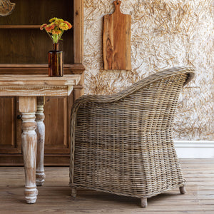 A rustic woven wicker armchair with a curved back and rolled arms, positioned next to a distressed wooden table in a styled room setting.