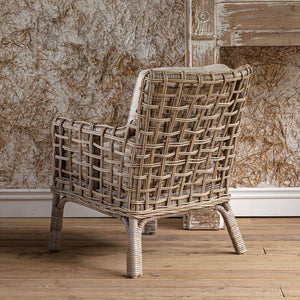 A woven rattan armchair with a distressed whitewashed finish, positioned on a wooden floor against a textured brown and beige wallpaper background.