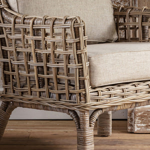 Close-up of a woven rattan outdoor chair with a light beige textured cushion, showing the detailed wicker construction and legs on a wooden floor.