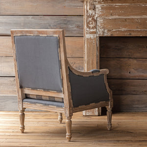 A rustic French-style armchair with a distressed brown wood frame and dark gray fabric upholstery, positioned in front of a weathered wooden wall and fireplace.