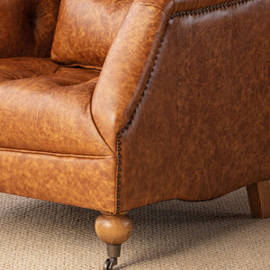 Close-up of a brown distressed leather armchair with button tufting, nailhead trim, and turned wooden legs on a textured beige carpet.