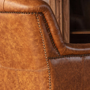 Close-up of a rustic brown leather armchair showing the textured grain, curved armrest, and dark metal nailhead trim.
