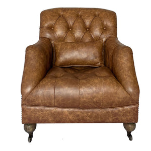 A brown tufted leather armchair with a matching cushion, shown against a plain white background. The chair features distressed leather upholstery, button tufting on the backrest and seat, rolled arms, and dark turned legs with casters.