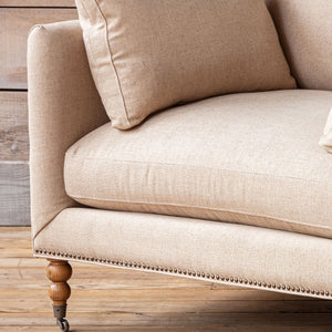 Close-up of a beige upholstered sofa corner with a matching cushion, showing the fabric texture, nailhead trim, wooden leg, and rustic wood floor and wall background.