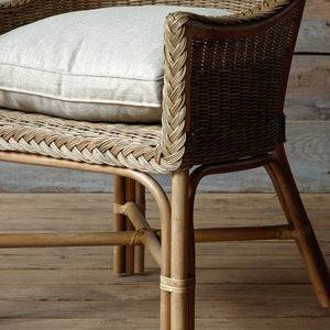 Close-up of a woven rattan chair with a light beige cushion, showing the wicker backrest, seat, and natural wood legs on a wooden floor.