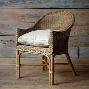 A natural woven rattan armchair with a curved back, rope-wrapped arm details, and a light beige cushion, set against a rustic wood plank wall and floor.