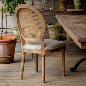 Wooden chair with wicker backrest and cushioned seat in a rustic setting with a wooden table and potted plants. Rosemere Round Cane-Back Dining Chair