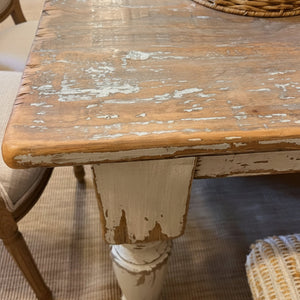 A rustic distressed wooden dining table with a weathered brown top and a white-painted pedestal leg, shown in a home setting with a woven basket and chair visible.