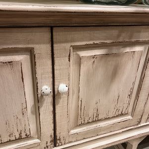 A close-up of a distressed light beige wood cabinet with two white knobs, showing a price tag hanging from one knob, in a furniture store setting.