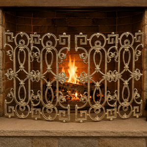 An ornate cream-colored metal fireplace screen with intricate scrollwork and Greek key motifs, standing in front of a brick fireplace with a burning log. A stone hearth sits below, flanked by a potted plant on the left and a black metal fireplace tool set on the right.
