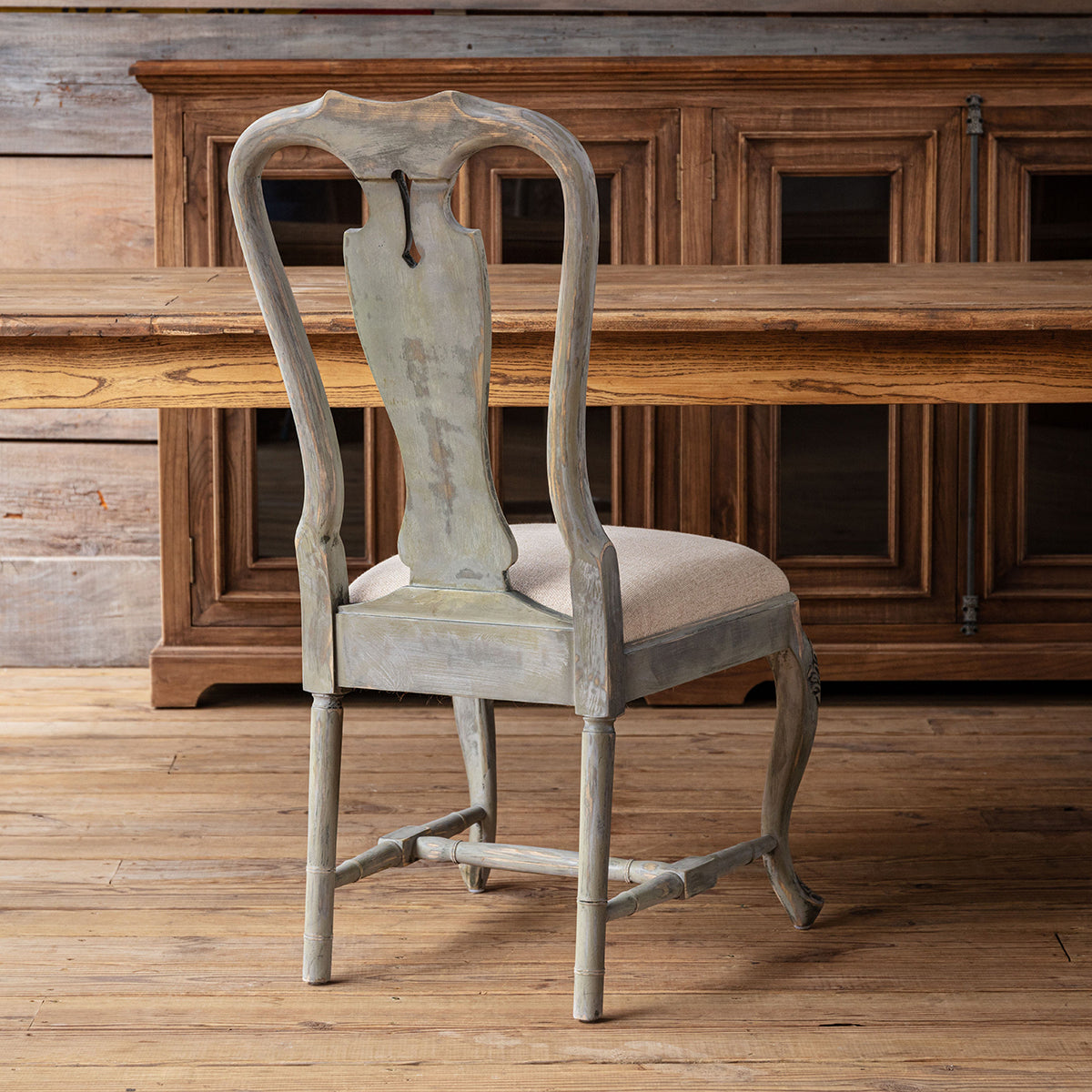 A distressed gray wooden dining chair with a light beige upholstered seat, positioned in front of a rustic wooden cabinet and table on a wooden floor.