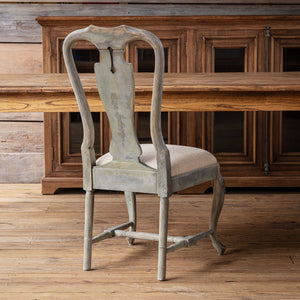 A distressed light gray wooden dining chair with a light beige upholstered seat, positioned in front of a rustic wooden dining table and matching cabinet on a wooden floor.