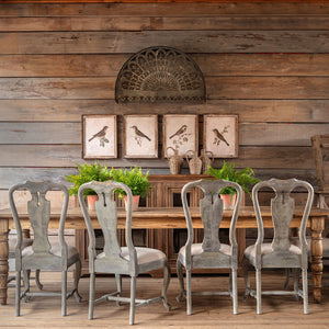 Four distressed gray wooden dining chairs arranged around a rustic wooden table in a styled dining room with a wood plank wall, bird prints, and potted green plants.