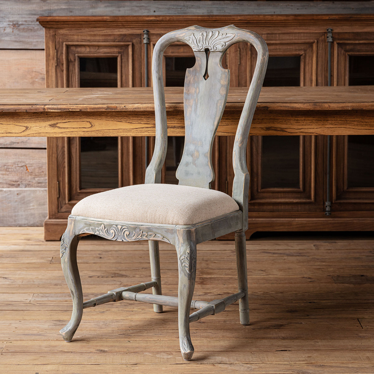 A distressed gray wooden dining chair with a light beige upholstered seat, positioned in front of a rustic wooden cabinet and table on a wooden floor.