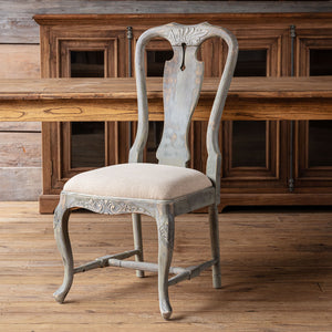 A distressed gray wooden dining chair with a light beige upholstered seat, positioned in front of a rustic wooden cabinet and table on a wooden floor.