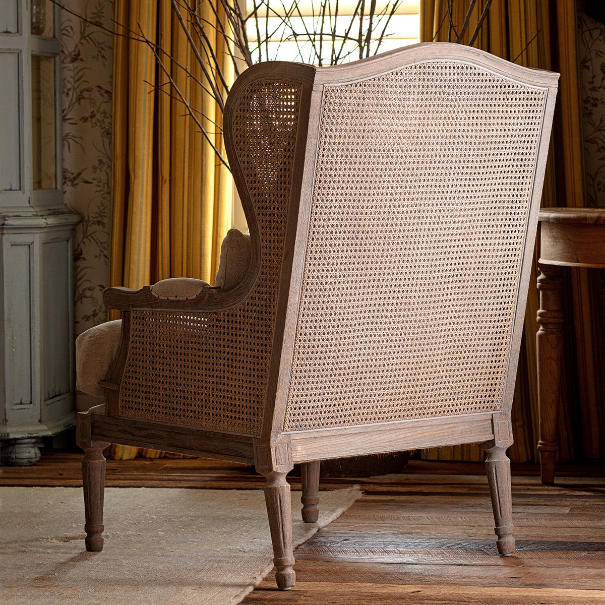 A rustic wingback armchair with a woven cane back and seat, upholstered in a light beige linen fabric, shown in a styled room setting with warm curtains and wooden flooring.