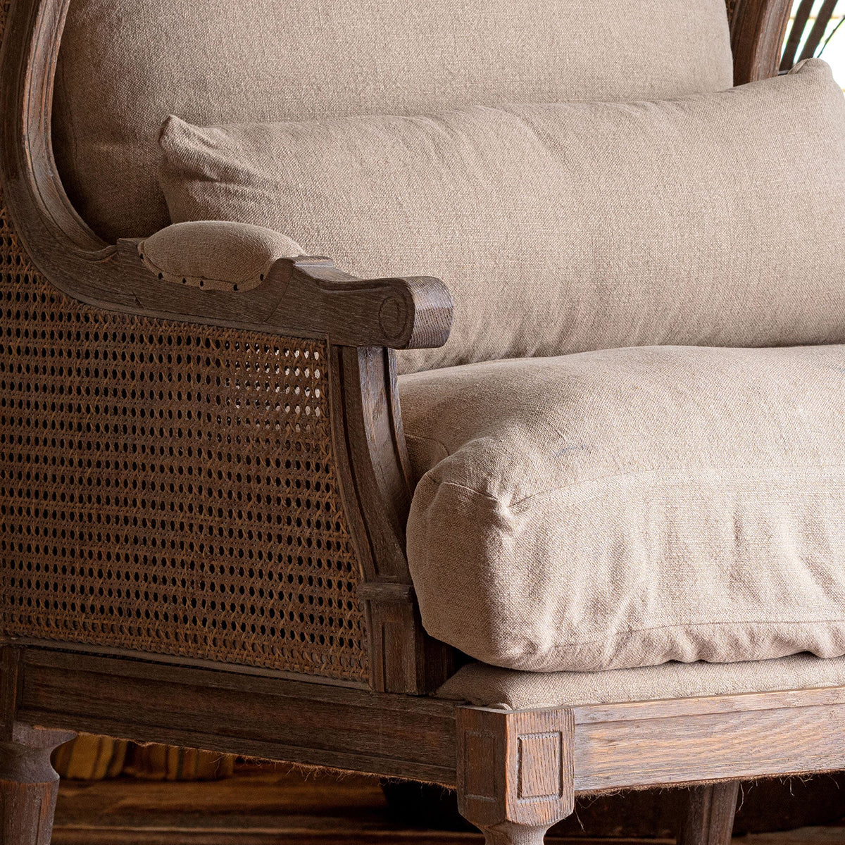 Close-up of a wooden armchair with woven cane panels and thick beige upholstered cushions.