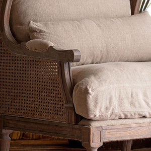 Close-up of a wooden armchair with woven cane panels and thick beige upholstered cushions.