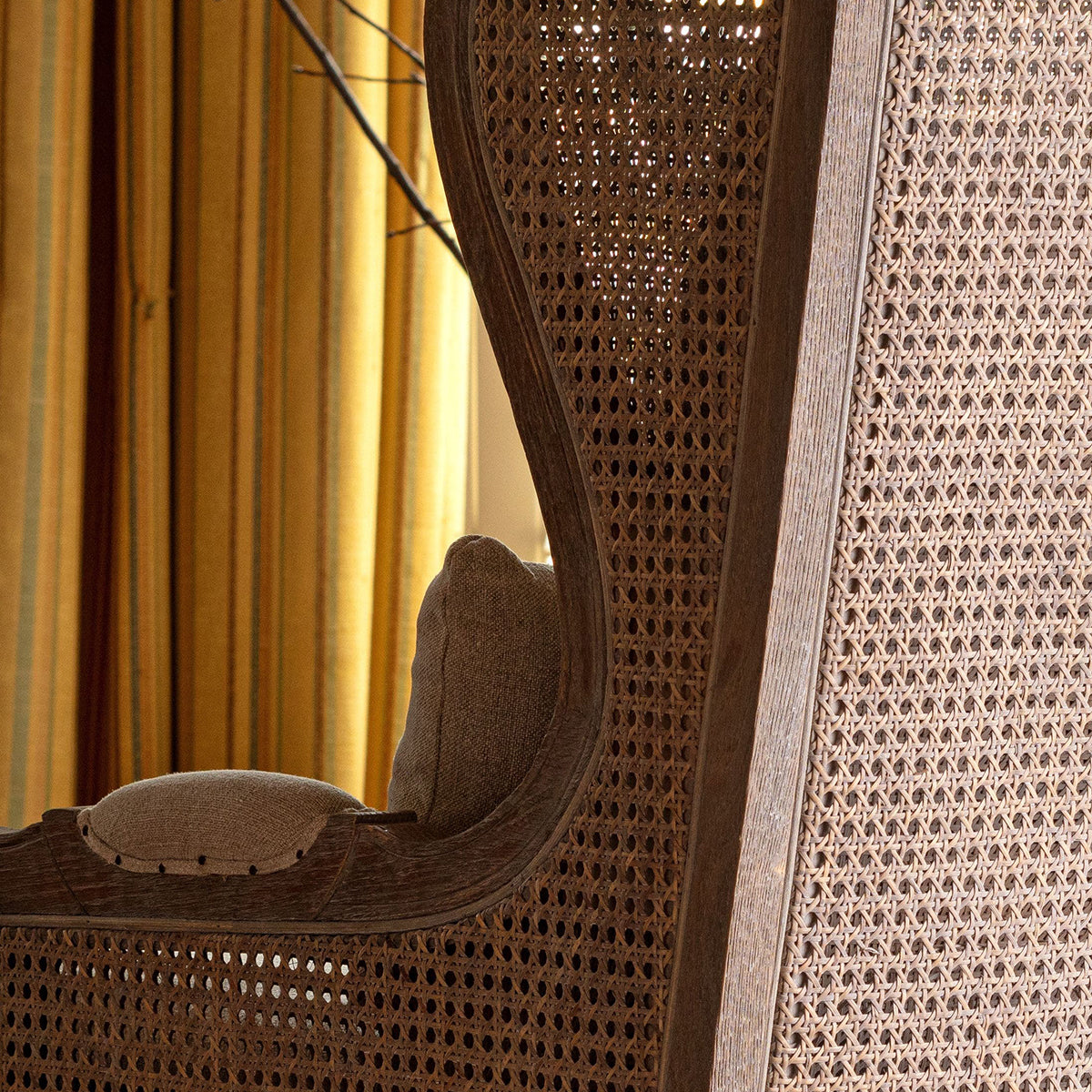A close-up of a woven cane or rattan chair back and armrest in warm brown tones, with a beige upholstered seat cushion and a softly lit indoor background with yellow curtains.