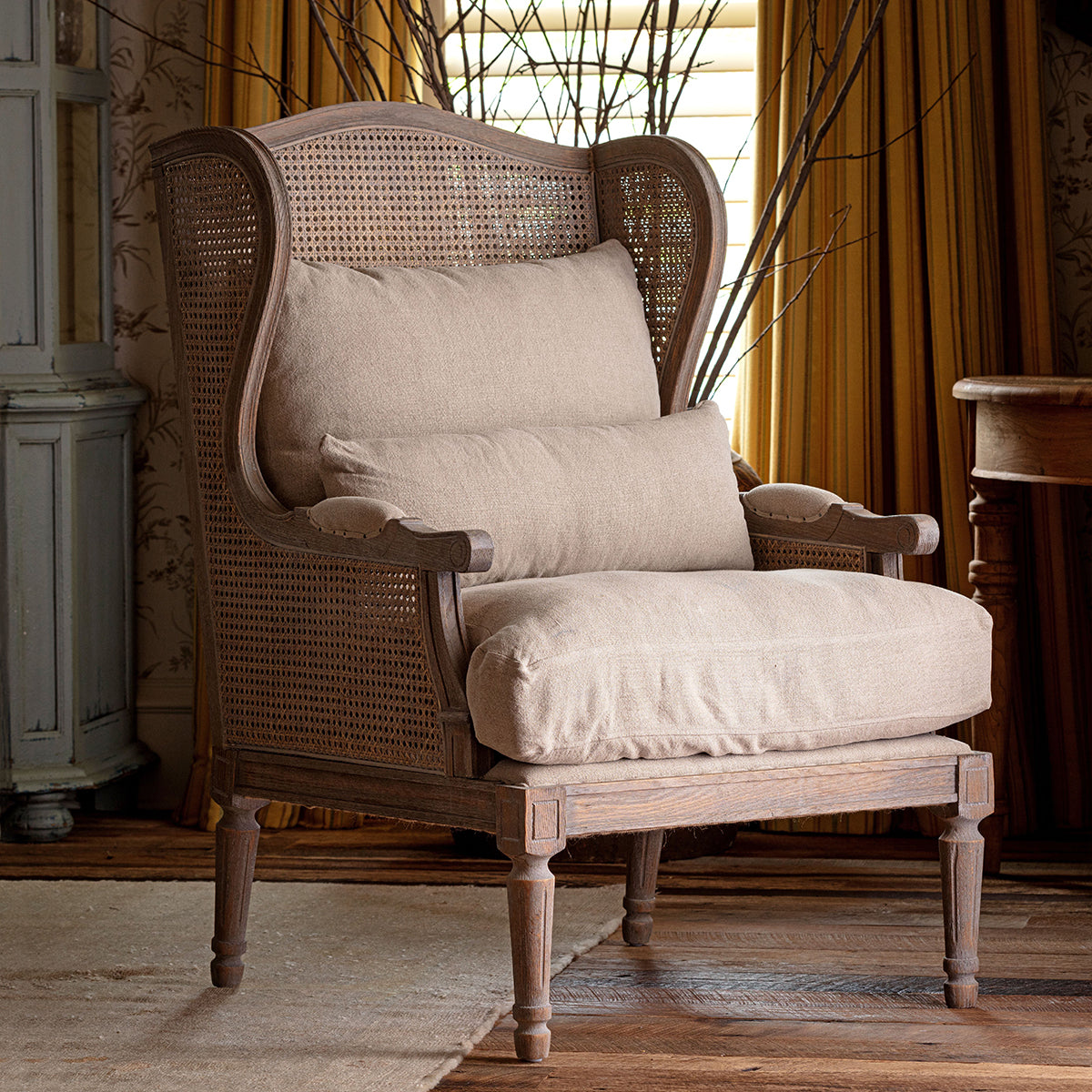 A rustic wingback armchair with a woven cane back and seat, upholstered in a light beige linen fabric, shown in a styled room setting with warm curtains and wooden flooring.