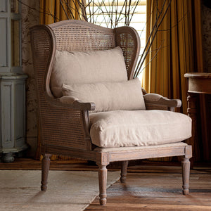 A rustic wingback armchair with a woven cane back and seat, upholstered in a light beige linen fabric, shown in a styled room setting with warm curtains and wooden flooring.