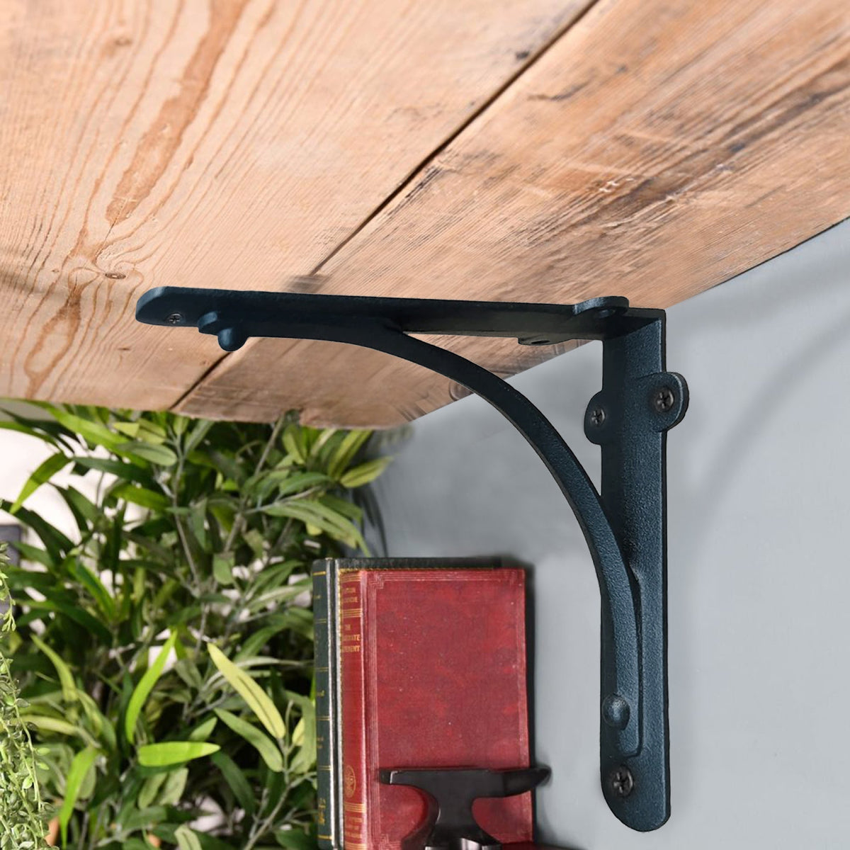 A black metal bracket shelf support mounted on a light gray wall beneath a rustic wooden tabletop, with green plants and a red book visible below.