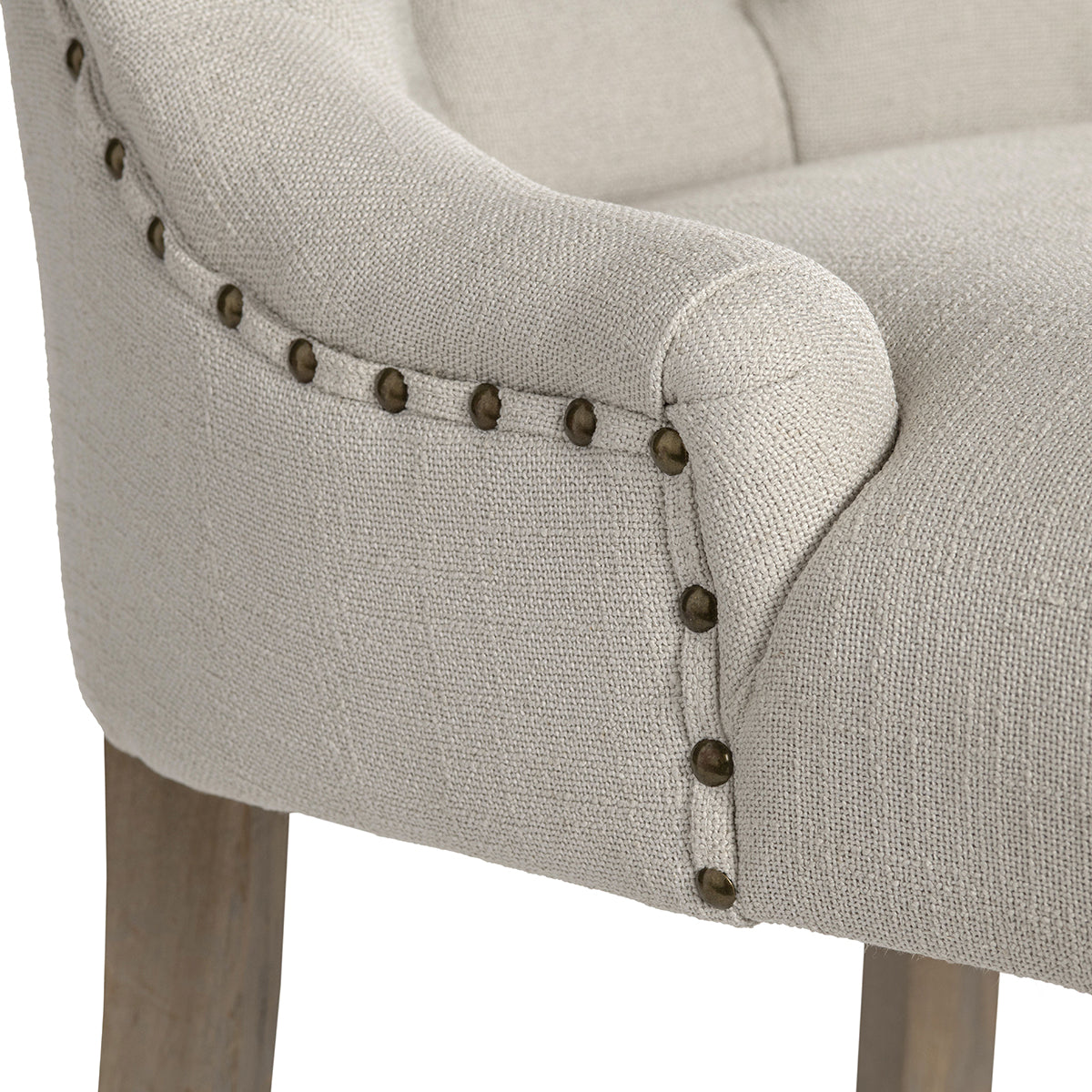 Close-up of a light beige upholstered armchair showing the fabric texture, rolled arm, nailhead trim, and part of the wooden leg against a white background.