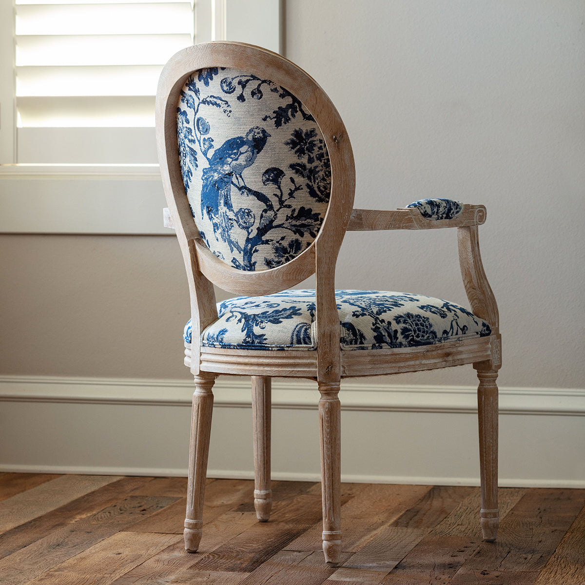 An antique-style wooden dining chair with a distressed light oak frame and blue and white toile-patterned upholstery, set on a rustic wooden floor in front of a window.