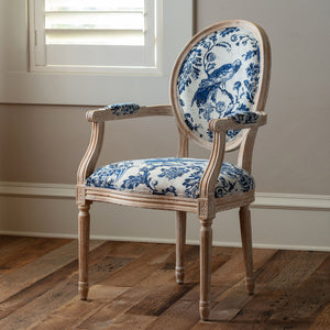 An antique-style wooden dining chair with a distressed light oak frame and blue and white toile-patterned upholstery, set on a rustic wooden floor in front of a window.
