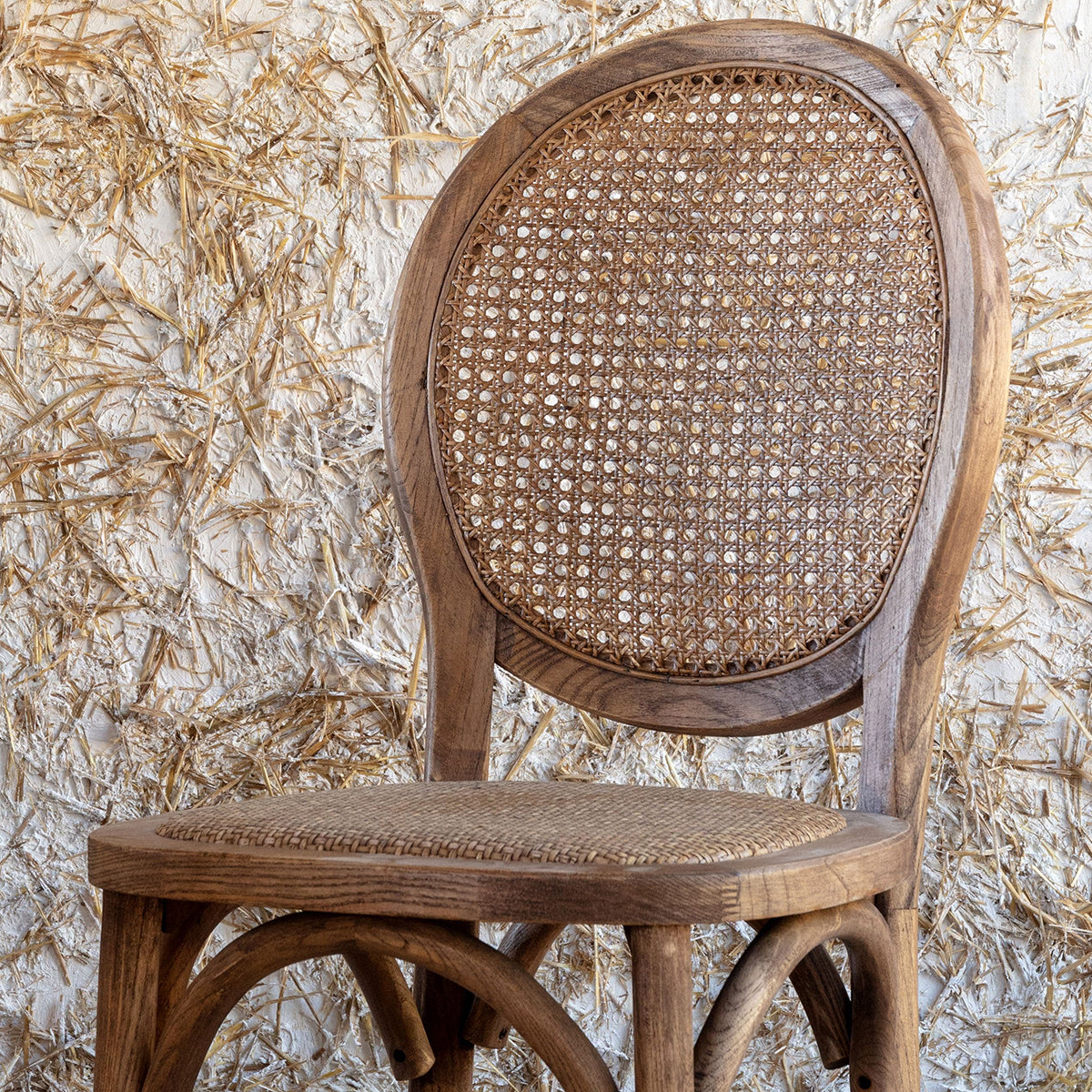 A rustic French bistro-style wooden bar stool with a woven cane backrest and seat, set against a textured beige wall and light wood floor.