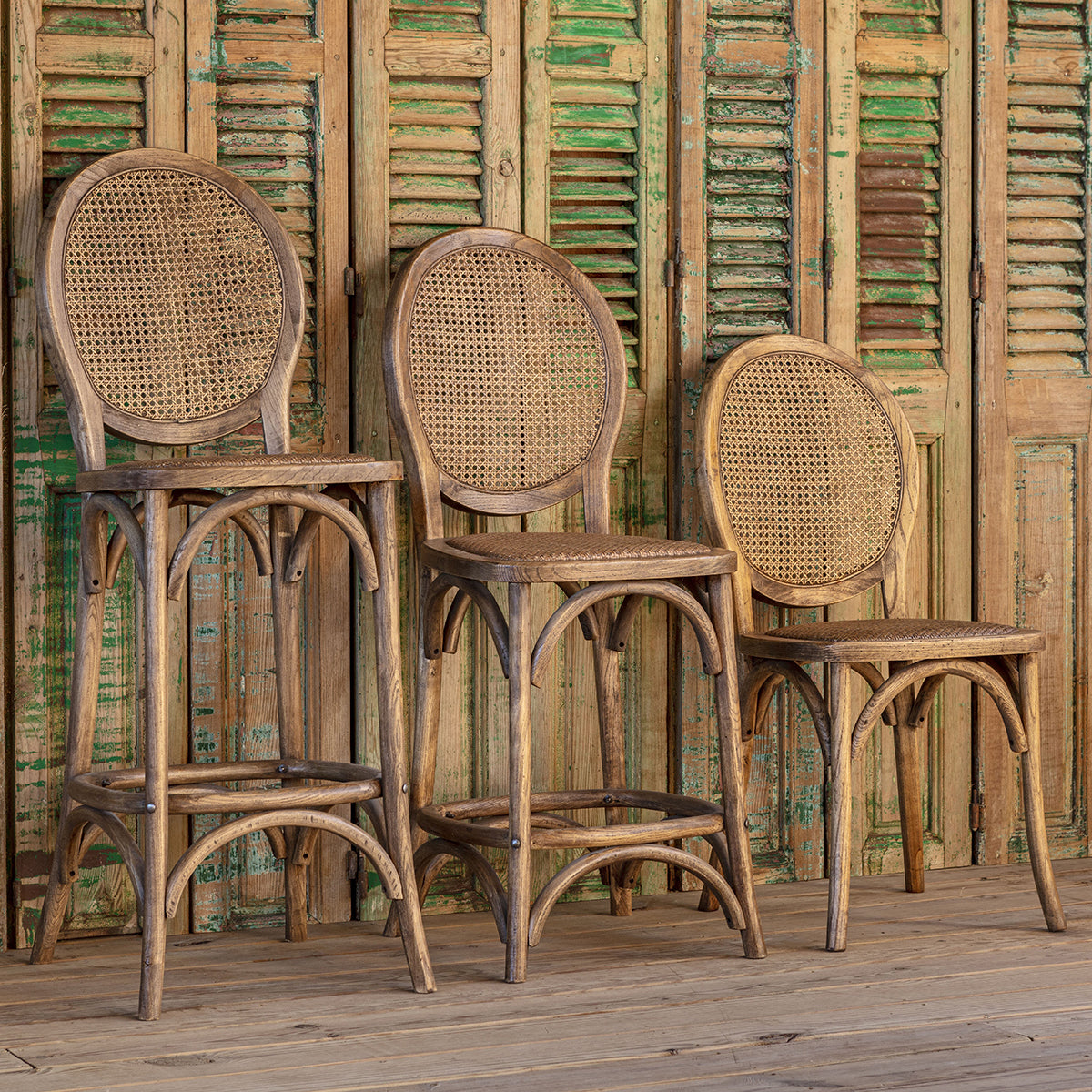 Three rustic wicker and bentwood bar stools with oval cane backs are arranged against a weathered green and brown wooden shutter backdrop.