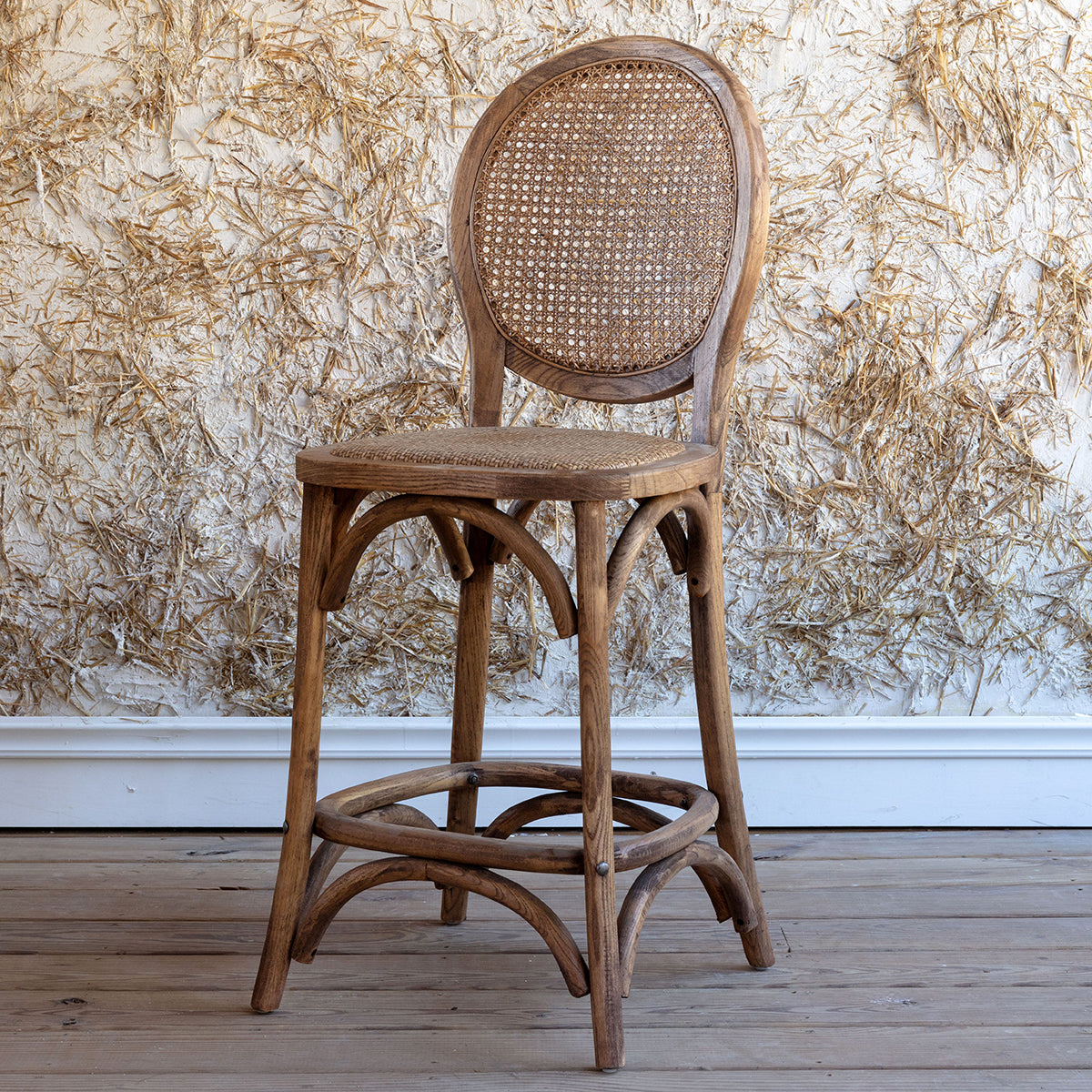 A rustic French bistro-style wooden bar stool with a woven cane backrest and seat, set against a textured beige wall and light wood floor.