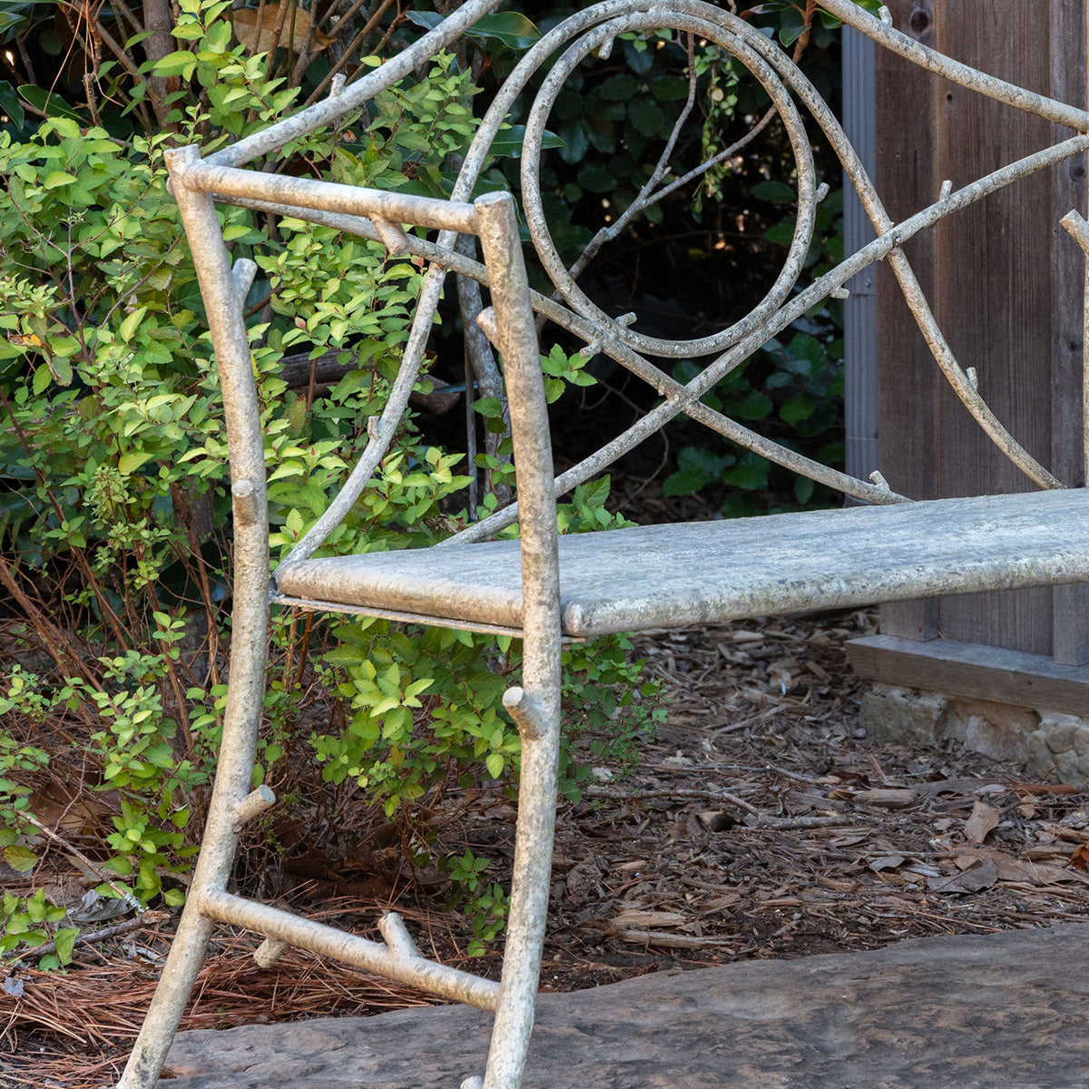 A rustic garden bench made from weathered branches and vines, set outdoors in a garden with green foliage, mulch, and a wooden fence in the background.