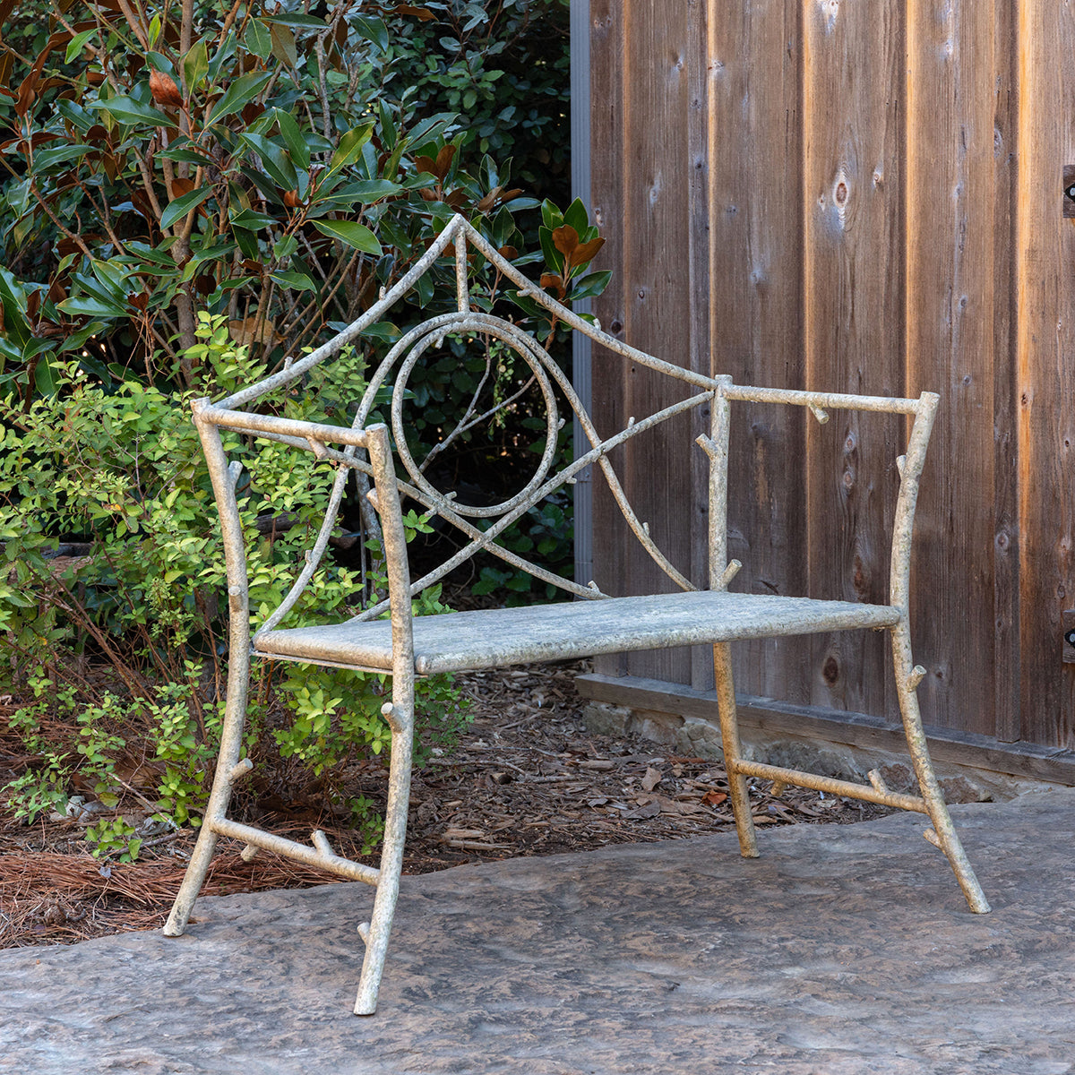 A rustic garden bench made from branches and metal, featuring a circular center motif and a weathered gray seat, set outdoors against a wooden fence and greenery.
