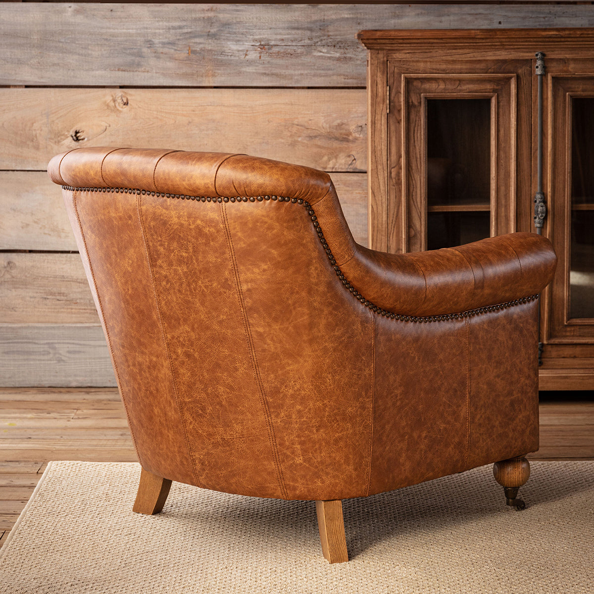 A brown leather armchair with a curved back, button tufting, nailhead trim, and wooden legs, shown in a rustic room setting with a wood-paneled wall and a wooden cabinet.