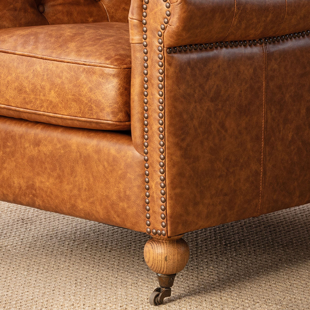 Close-up of a brown leather armchair corner with brass nailhead trim and a turned wood caster leg on a textured beige carpet.