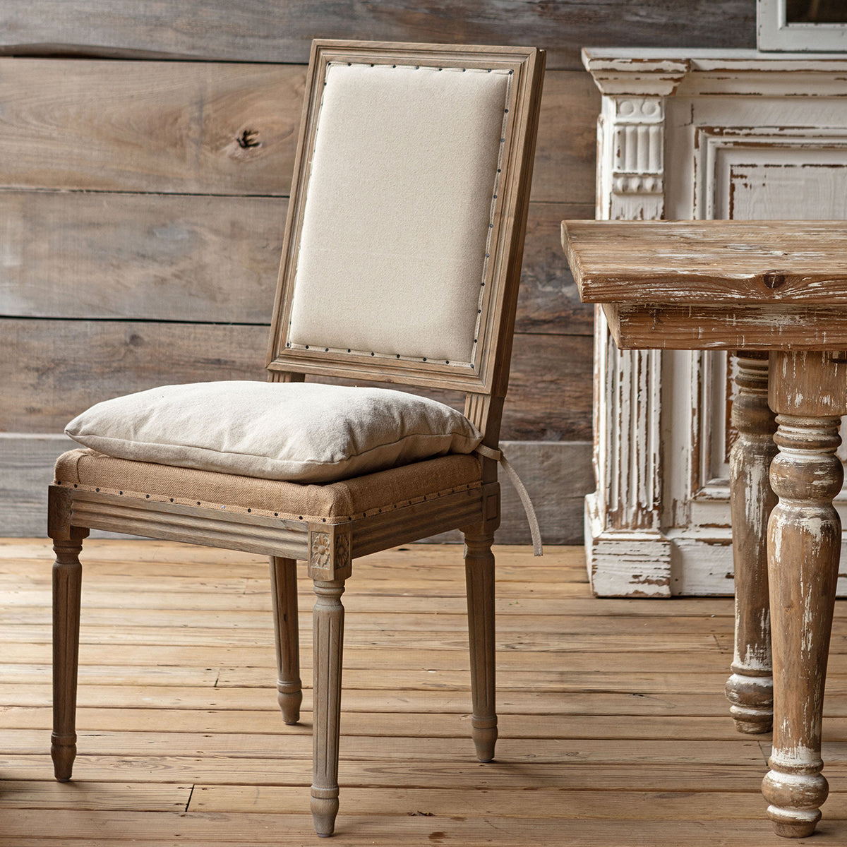 A rustic French-style dining chair with a distressed gray-brown wood frame, cream upholstered backrest, and a matching cushion on the seat, set in a rustic room with a wooden table and distressed furniture in the background.