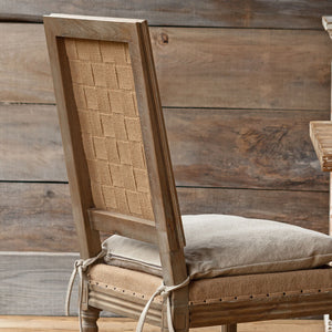 A rustic wooden dining chair with a woven cane backrest, upholstered in a warm brown fabric, and a light beige cushion tied with white strings, set against a rustic wood plank wall.