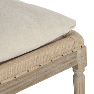 Close-up of a upholstered bench or ottoman corner, showing a light beige fabric cushion, woven tan upholstery, dark nailhead trim, and a weathered light wood frame on a white background.