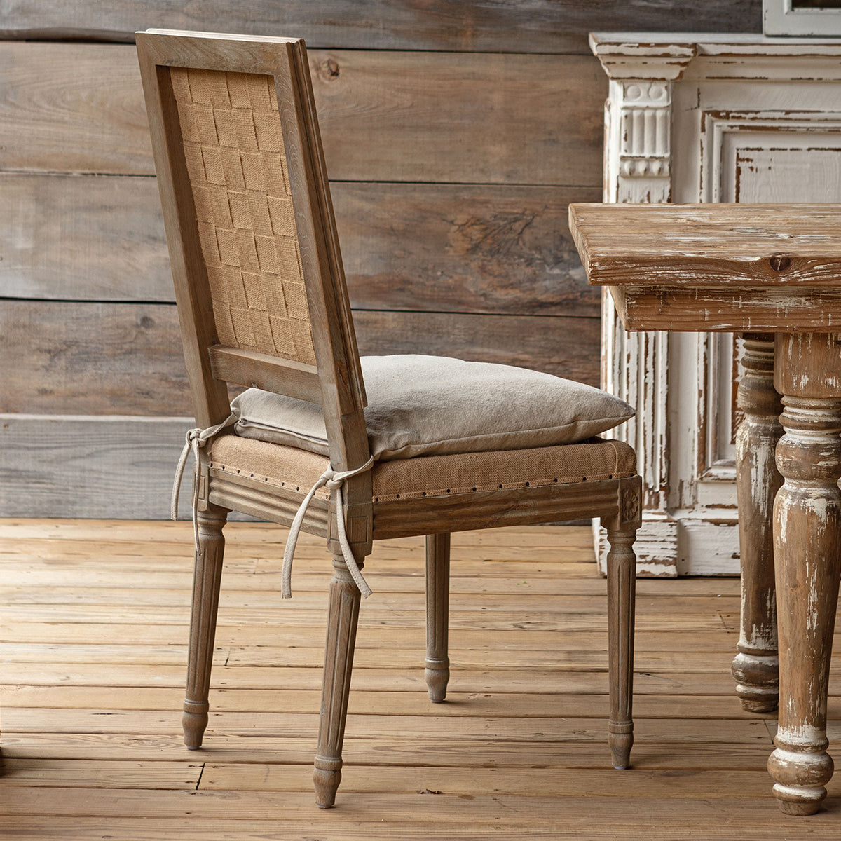 A rustic French-style dining chair with a distressed gray-brown wood frame, cream upholstered backrest, and a matching cushion on the seat, set in a rustic room with a wooden table and distressed furniture in the background.