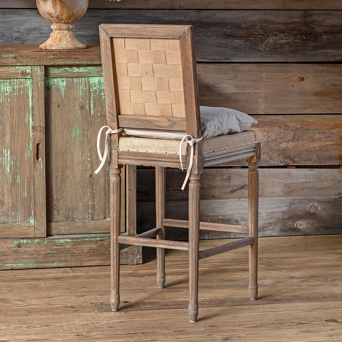 A rustic wooden barstool with a light beige upholstered backrest and seat cushion, styled in front of a distressed green cabinet against a wood-paneled wall.