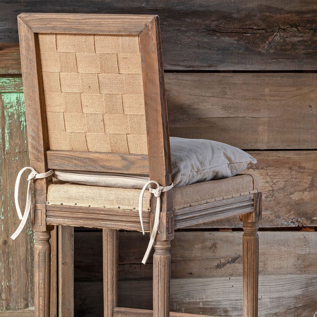 A rustic wooden chair with a woven burlap backrest, upholstered seat, and tied side ties, shown against a weathered wood plank background.