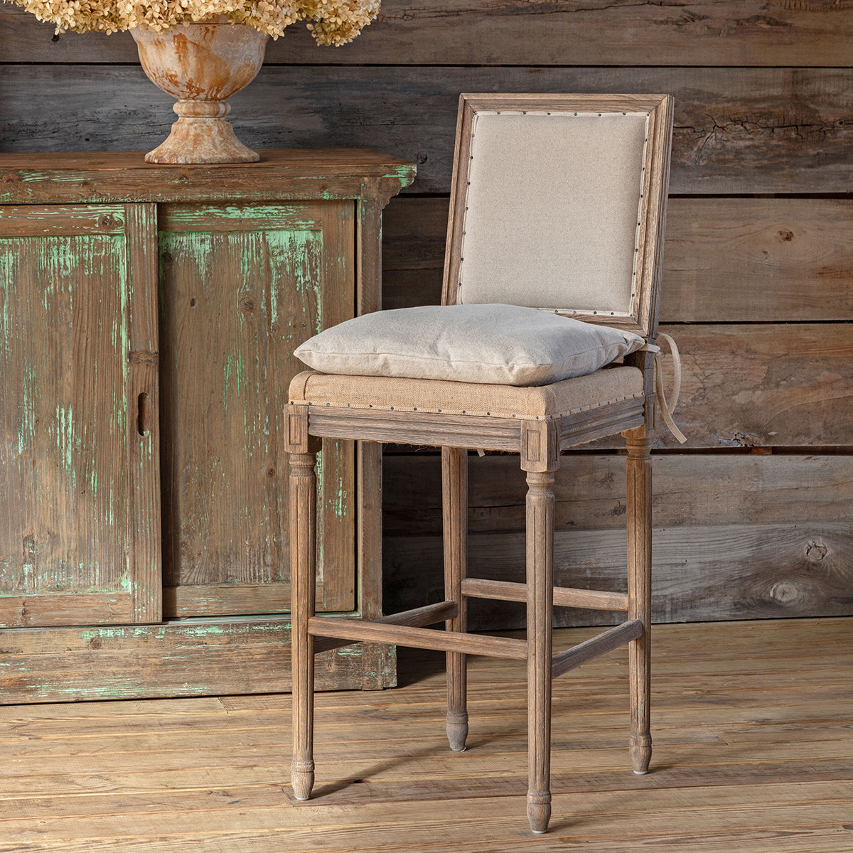 A rustic wooden barstool with a light beige upholstered backrest and seat cushion, styled in front of a distressed green cabinet against a wood-paneled wall.