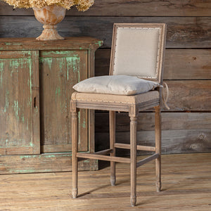 A rustic wooden barstool with a light beige upholstered backrest and seat cushion, styled in front of a distressed green cabinet against a wood-paneled wall.