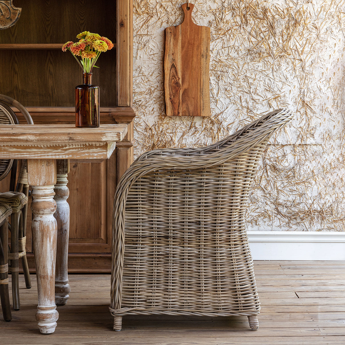 A woven wicker armchair with a light beige cushion sits beside a rustic wooden table in a styled interior setting with a textured wall and wooden decor.